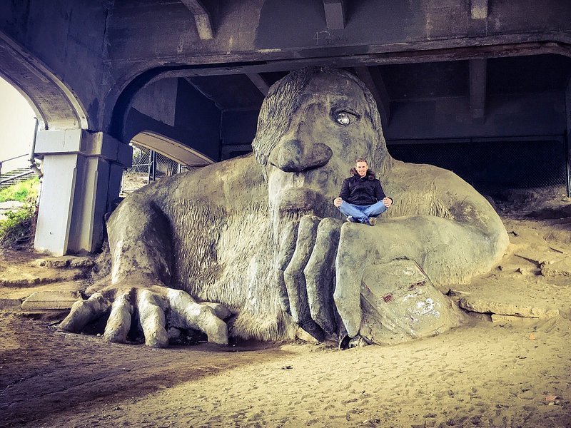 A large grey troll claws its way toward the camera with a tourist sitting on top of one of its hands