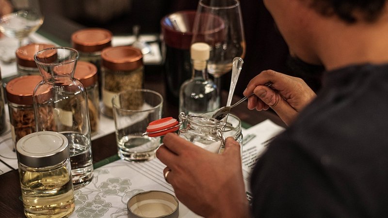 A person spoons sugar out of a jar and into a glass, surrounded by jars full of other ingredients