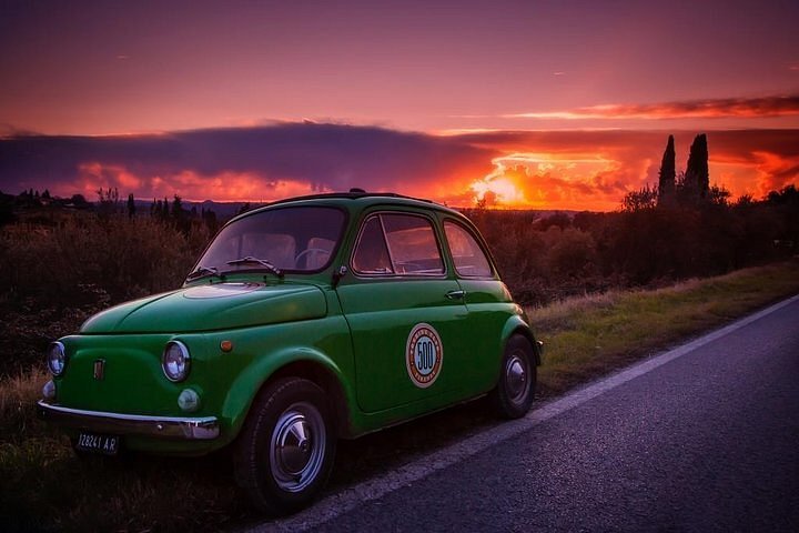 A green Fiat is parked on the side of the road during a purple-pink sunset