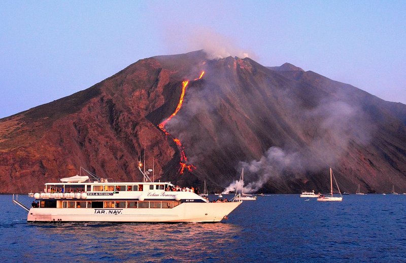 A boat sails in front of a volcano erupting with a thin strip of lava that cascades into the sea