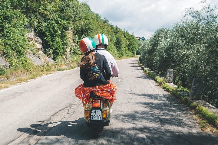 Two people ride a vespa on a narrow road surrounded by greenery