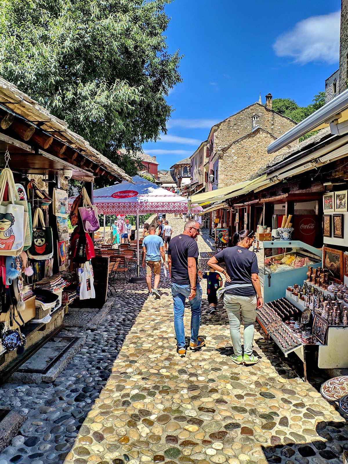 A narrow, bustling street in Mostar's Old Town with traditional shops displaying colorful souvenirs and crafts, under a warm light.