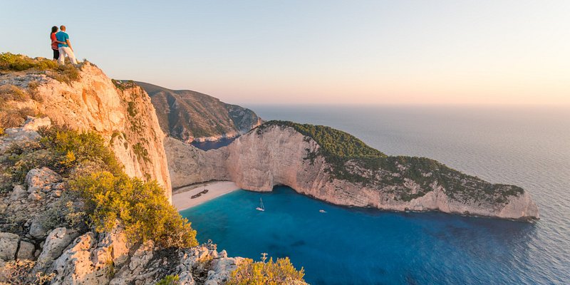 Couple enjoying sunset from a high cliff over famous Navagio shipwreck beach. Zakynthos, Greek Islands, Greece