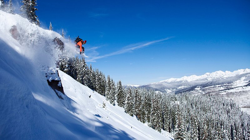 A snowboarder jumping off a cliff on a sunny powder day in Vail, Colorado 