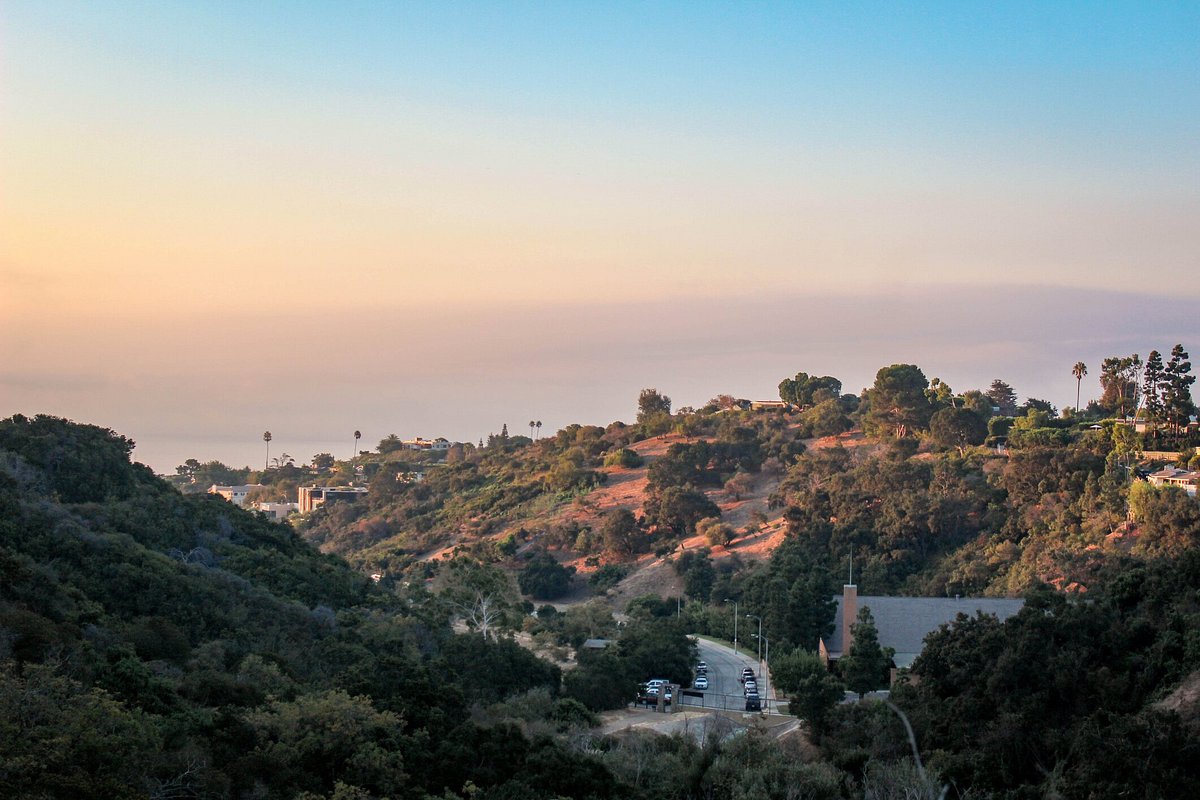 A concrete street with cars from a mountain top with green mountain ranges and houses, the ocean in the distance and the sky in hazy orange and blue during a California sunrise
