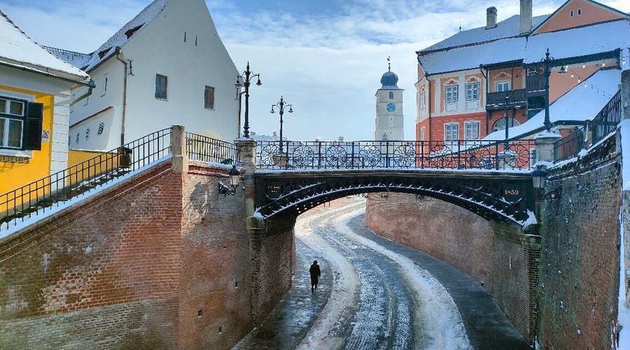 Pedestrian crossing the Bridge of Lies in Sibiu, Romania, with historic buildings and blue sky.