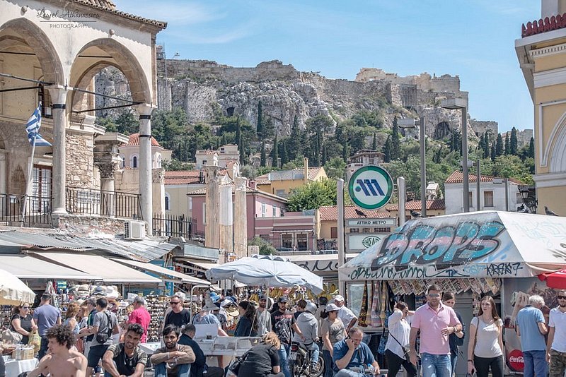 Monastiraki flear market vendors offer up goods to a crowd under individual canopies, with near modern buildings and ancient wall structures in the background