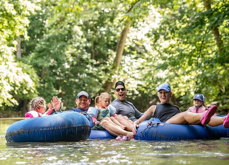 Group of adults and children on blue inner tubes floating down creek surrounded by trees