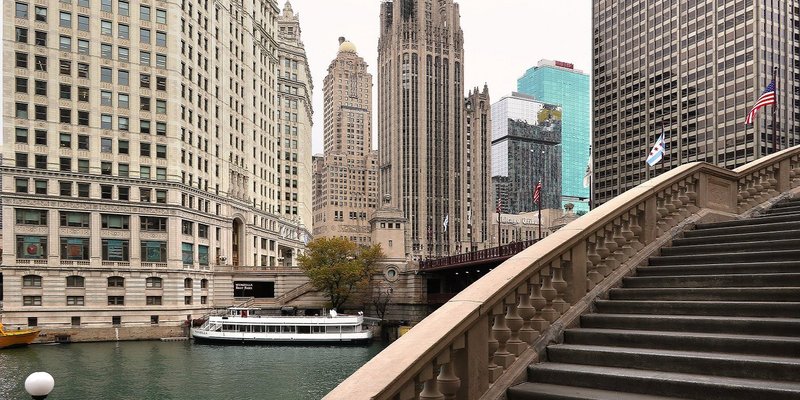 Boat docked in river, which is flanked by walkway and high-rise buildings.