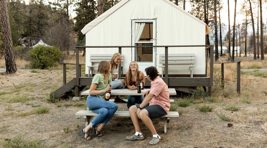 Four friends sharing a beverage at the picnic table in front of their cozy canvas cabin during golden hour. 