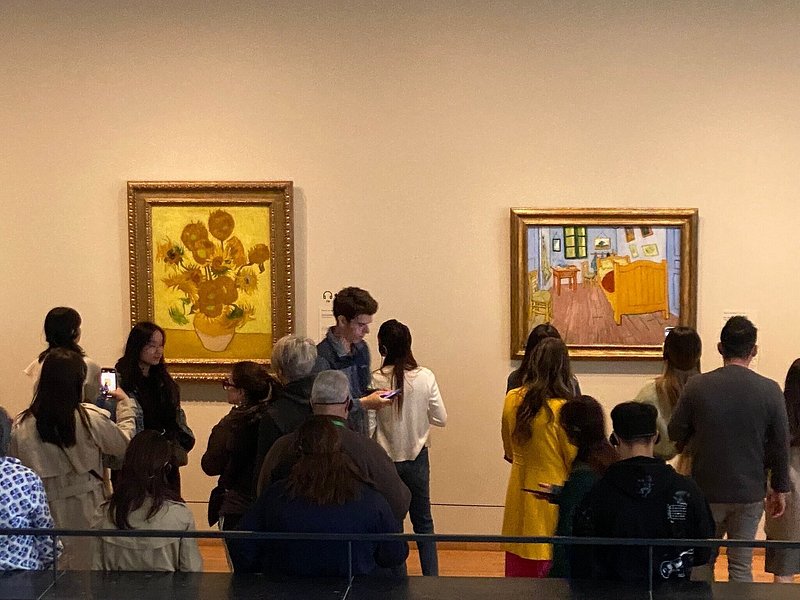 People walking around museum next to Van Gogh's Sunflowers and Bedroom in Arles paintings