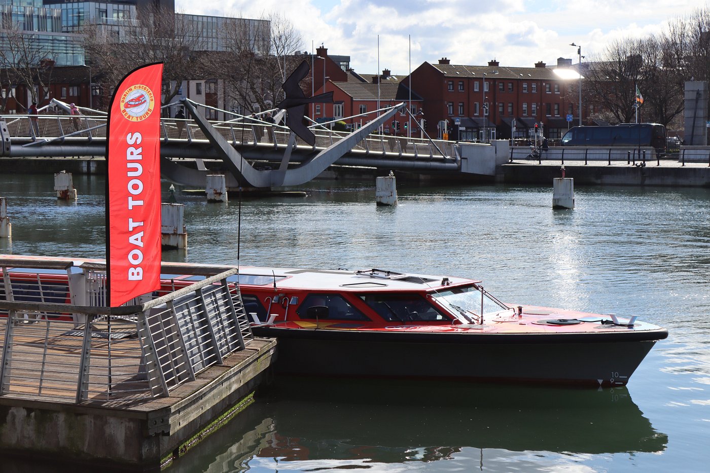 Red boat docked near a 'Boat Tours' sign by a river with a bridge.