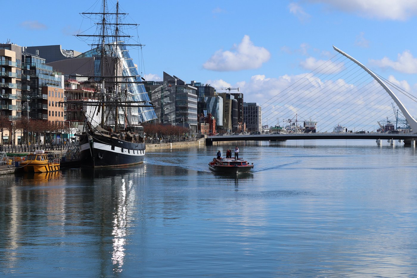 River scene with a tall ship, modern buildings, and a bridge under a blue sky.
