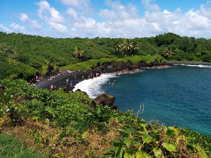 Vista panorâmica de viajantes a relaxar na praia de areia preta de Honokalani, em Maui, rodeada de vegetação exuberante e águas azuis cristalinas