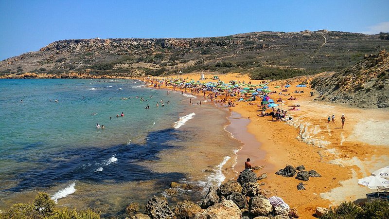 Vista aérea de viajantes a nadar e descontrair na Baía de Ramla, uma praia longínqua de areia laranja, rodeada por montanhas rochosas