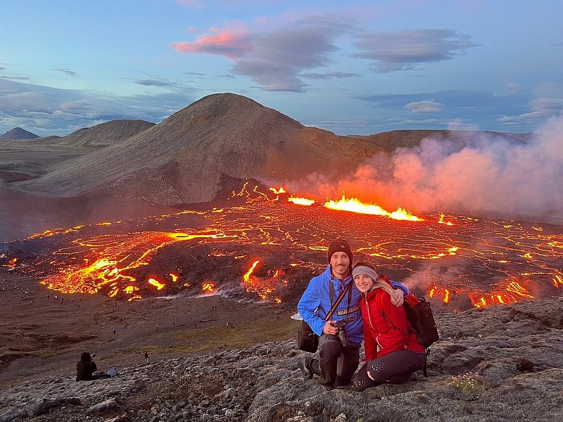 멀리 용암이 화산을 타고 흐르는 바위 풍경 위에서 포즈를 취하고 있는 두 사람