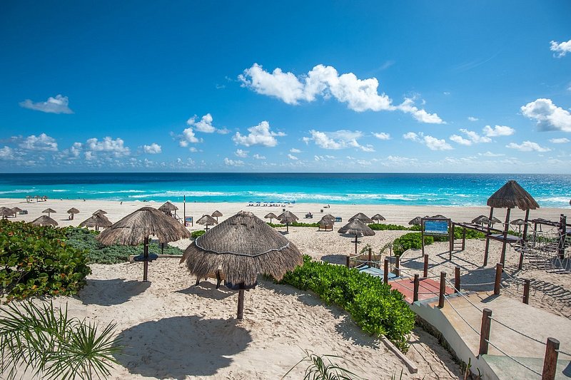 Beach with bright-blue water, and large sandy stretch lined with straw umbrellas