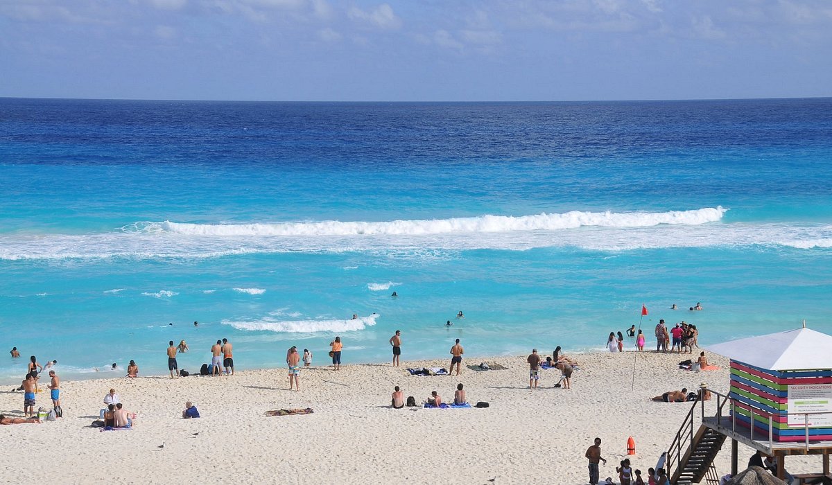 맑고 푸른 바닷물이 보이고 모래사장에 사람들이 서 있거나 앉아 있으며 인명구조 요원이 있는 해변