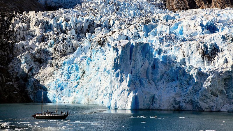 A sailboat surrounded by small icebergs floats in front of a glacier as it meets the ocean