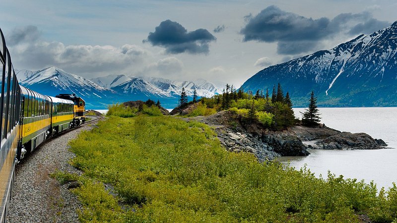 A green and yellow train snakes along a lake surrounded by snow-capped mountains