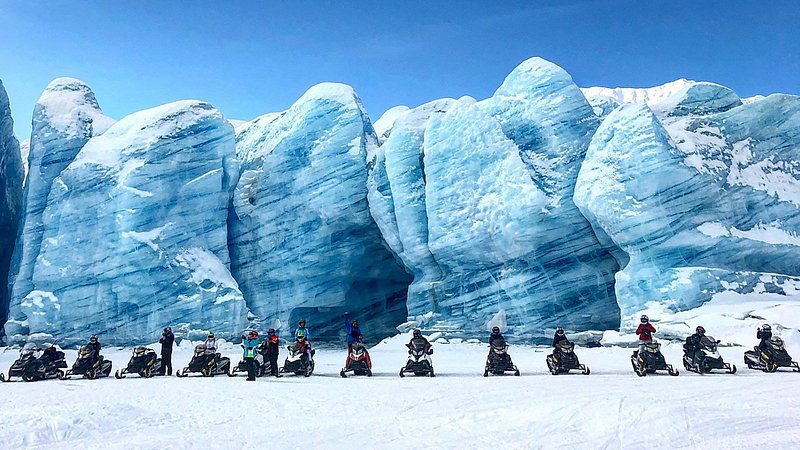 A line of people on snowmobiles in front of a ice-blue striated glacier