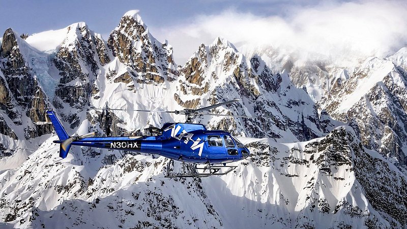 A blue helicopter flies in front of snow-covered, sheer mountain faces