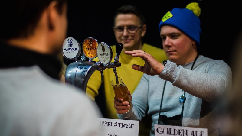 Two gentlemen stand next to each other while one pours beer from a tap