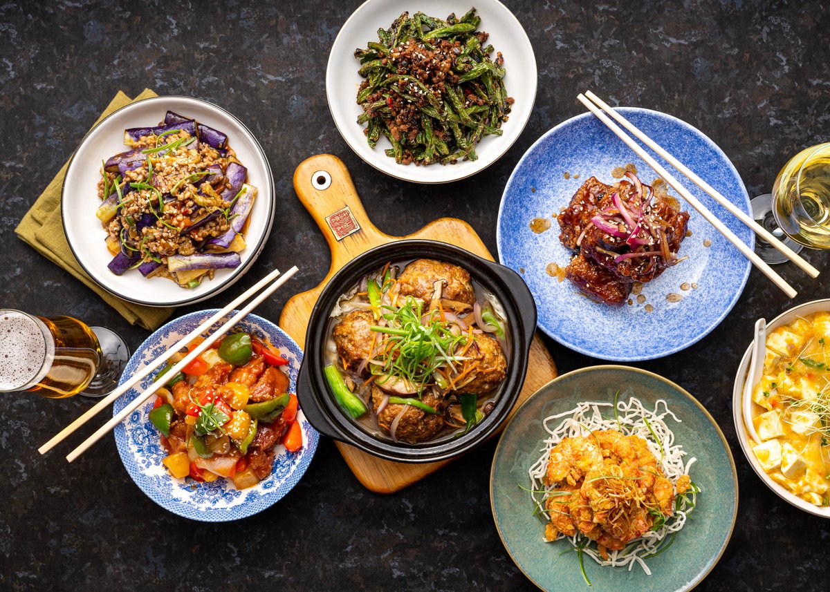 A top-view image showcasing a colorful spread of Chinese dishes on a dark tabletop. The variety includes stir-fried eggplant with minced meat, crispy green beans with sesame seeds, glazed pork ribs, sweet and sour chicken, a hotpot-style dish garnished with green onions, golden-fried shrimp on crispy noodles, and a tofu dish in a creamy sauce. Each dish is served on unique plates with chopsticks, accompanied by beverages such as beer and white wine, creating an inviting and vibrant dining setup. 