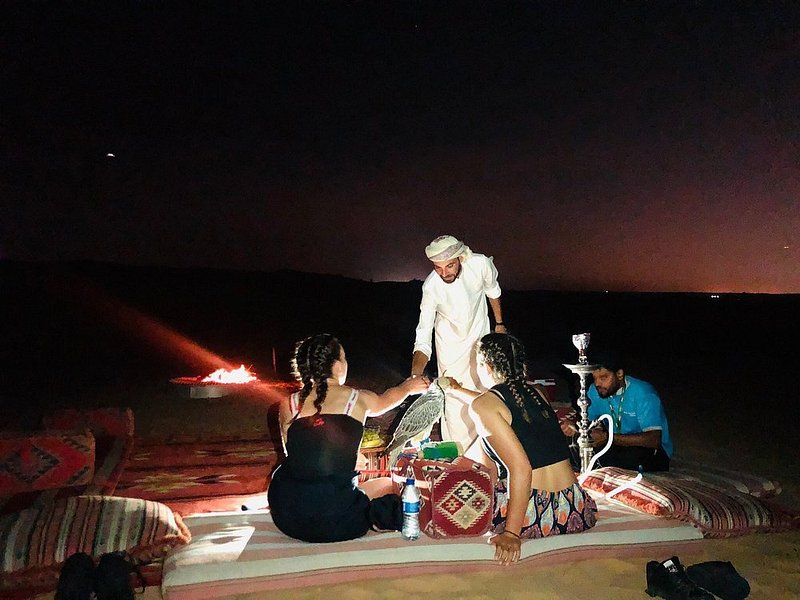 Twee reizigers die met hun gids ontspannen op kussens bij een kampvuur en genieten van de nachtelijke hemel boven de woestijn van Dubai.