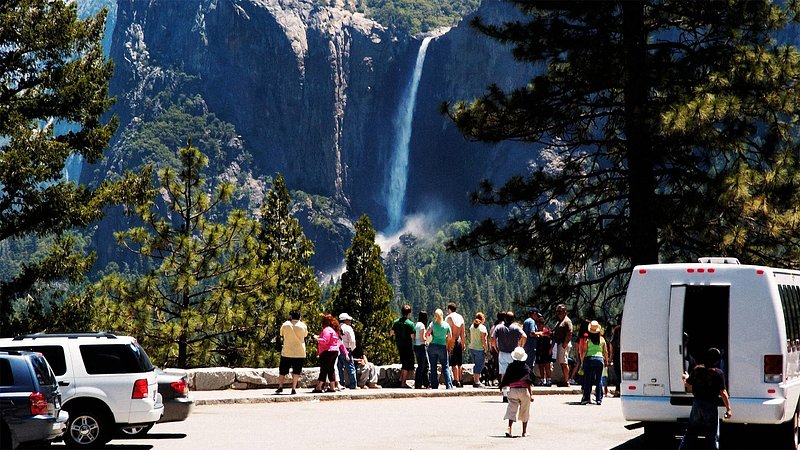 A group of tourists stand looking out at a waterfall cascading down a snow-capped mountain