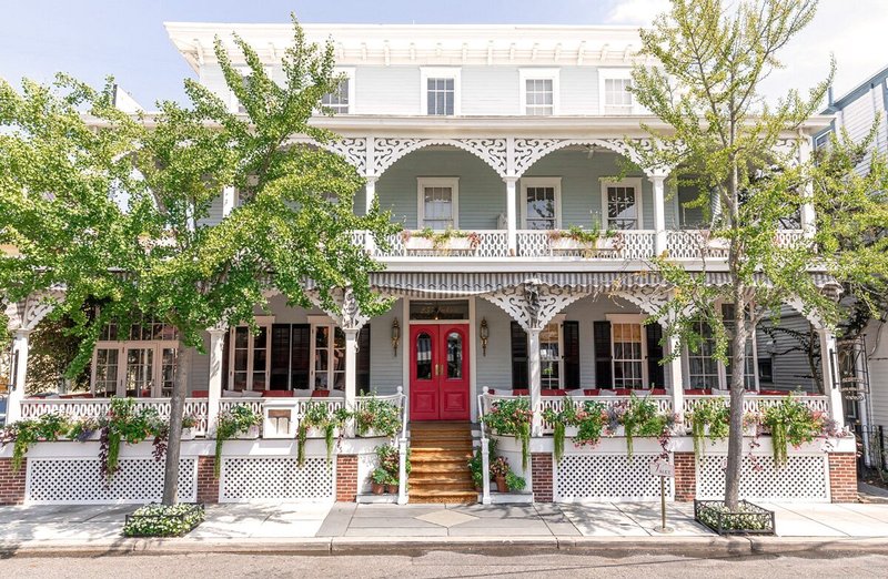 Exterior of three-story, light-blue inn with red door and lots of plants along porch railing