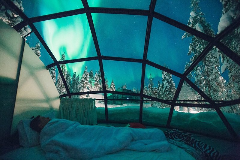 A male traveler laying in bed in a glass igloo, gazing up at the Northern Lights