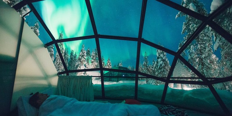 A male traveler laying in bed in a glass igloo, gazing up at the Northern Lights.
