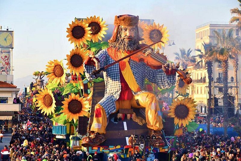 Πλήθος κόσμου κοιτάζει ένα τεράστιο άρμα που απεικονίζει έναν άνδρα, περιτριγυρισμένο από ηλιοτρόπια, να παίζει βιολί στο καρναβάλι του Βιαρέτζιο στην Ιταλία