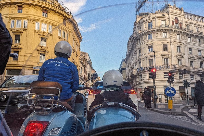 A man is bringing a tourist around Rome in a Vespa. The man is on the scooter, while the tourist is seated in its sidecar.