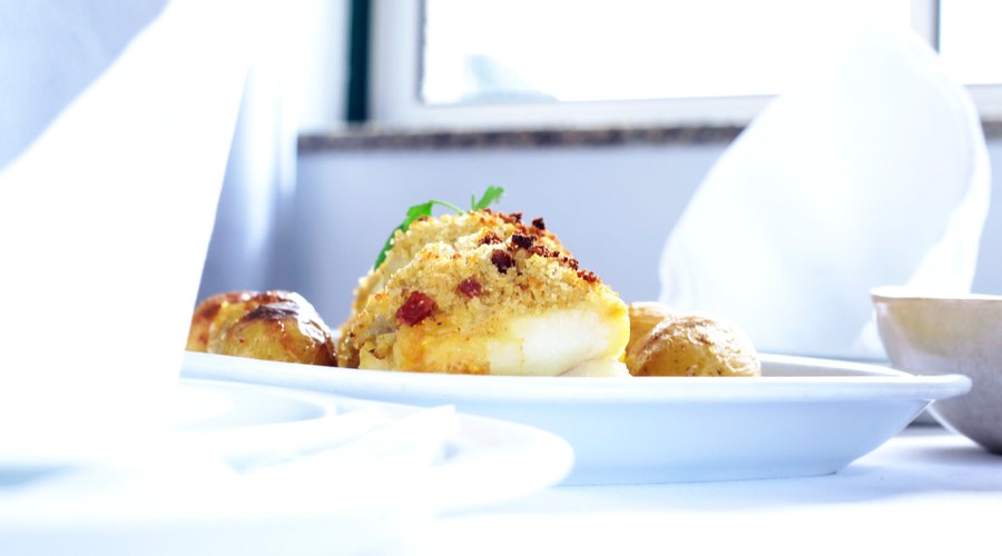  A deliciosa tradição portuguesa ganha destaque nesta elegante apresentação do Miracastro Restaurante, situado na pitoresca vila de Castro Laboreiro. À vista, temos um bacalhau dourado, coberto com uma crosta de broa crocante, realçado por um fio de azeite extra virgem que se espalha pela cerâmica branca do prato. Ladeando o peixe, pequenas batatas ao murro compartilham do azeite, adicionando uma textura rústica ao conjunto.