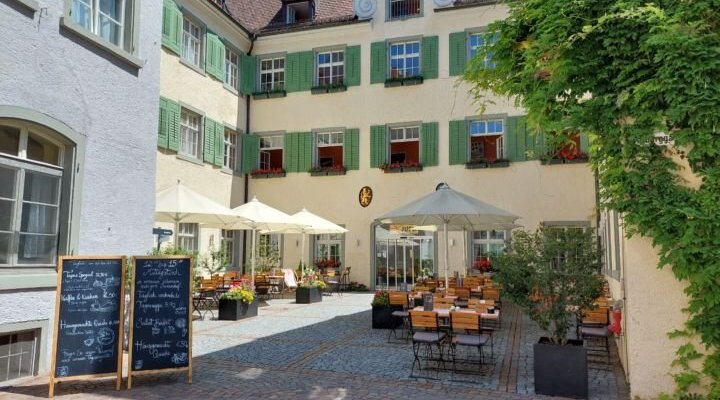 Sie sehen die Sonnenterrasse mit Tischen, Stühlen und Sonnenschirmen im Innenhof im JUFA Hotel Meersburg am Bodensee in Baden-Württemberg in Deutschland.