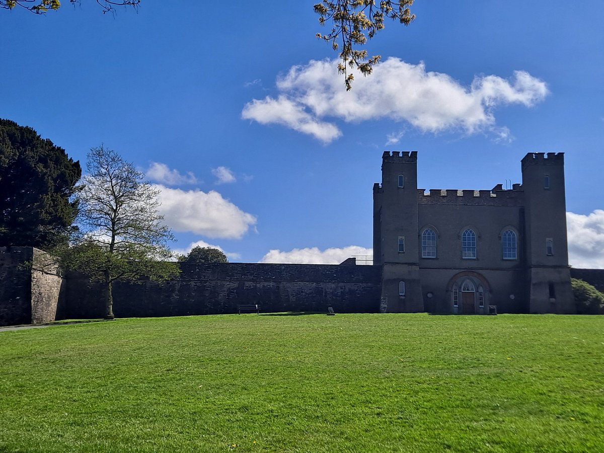 The historic Hillsborough Fort in Hillsborough, Northern Ireland, showing its imposing stone walls and entrance, standing amidst green grass under a partly cloudy sky.
