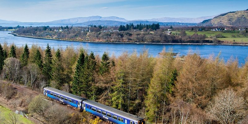 A West Highland Line on its journey through forests next to a river.