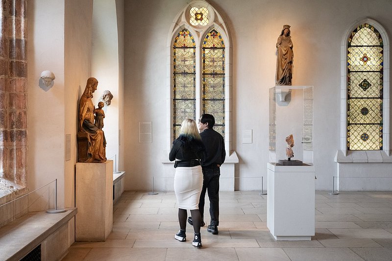 Two people viewing sculptures and stained-glass windows within Met Cloisters