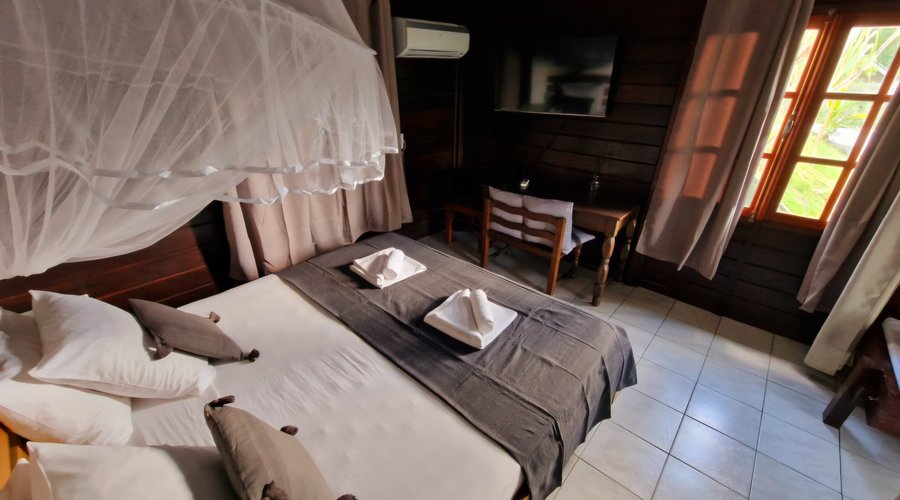 Votre cocon tropical à Deshaies Chambre alliant charme créole et confort moderne, idéale pour se ressourcer. Profitez d’un lit double avec moustiquaire, climatisation, TV écran plat et coin bureau, le tout baigné de lumière naturelle grâce aux grandes fenêtres. Le cadre chaleureux et le mobilier en bois offrent une ambiance relaxante, parfaite pour un séjour mémorable en Guadeloupe.