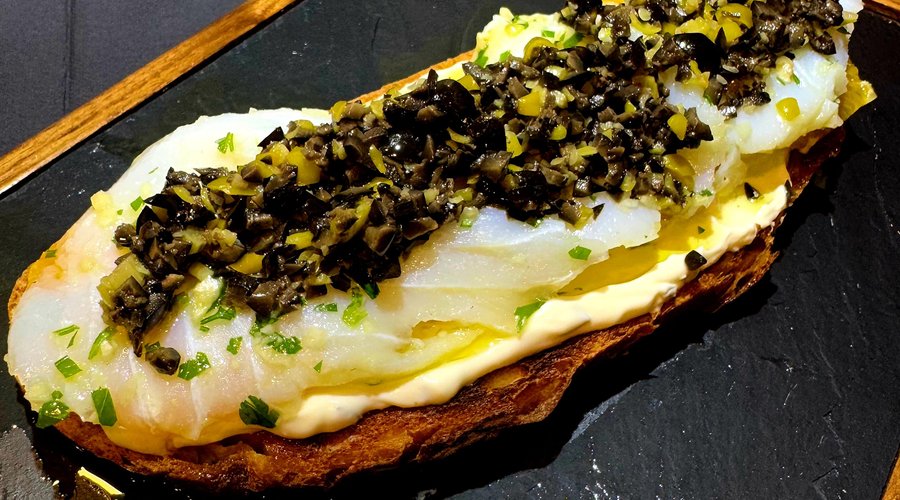 Tapa de la semana del 10 al 15 de Marzo de 2025. Pan tostado masa madre, salsa tartara, rodajas de naranja natural, bacalao fresco macerado con vinagreta de guindilla picante y aceitunas negras.