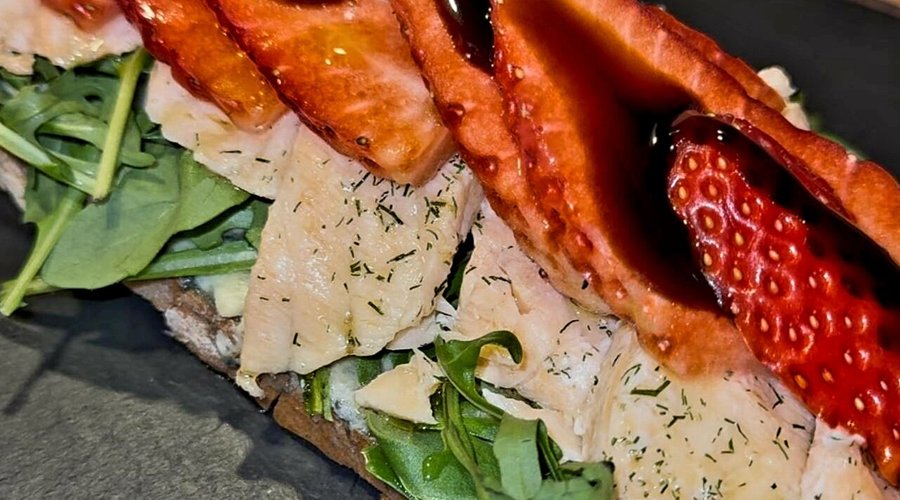 Tapa de la semana del 17 al 22 de Marzo de 2025. Pan tostado masa madre, queso azul danés, rucula fresca selvática, salmón fresco al horno, láminas de fresas y reducción de modena.