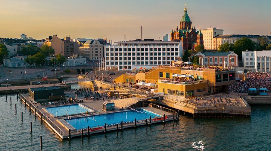 A modern outdoor swimming complex, Allas Sea Pool, with saunas and pools, situated by the waterfront in Helsinki, Finland, with city buildings in the background.