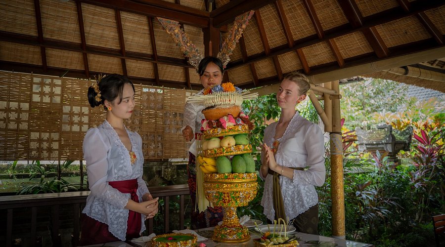 Three women dressed in traditional Balinese attire are arranging a tall and colorful gebogan offering made of fruits and flowers under a thatched pavilion.